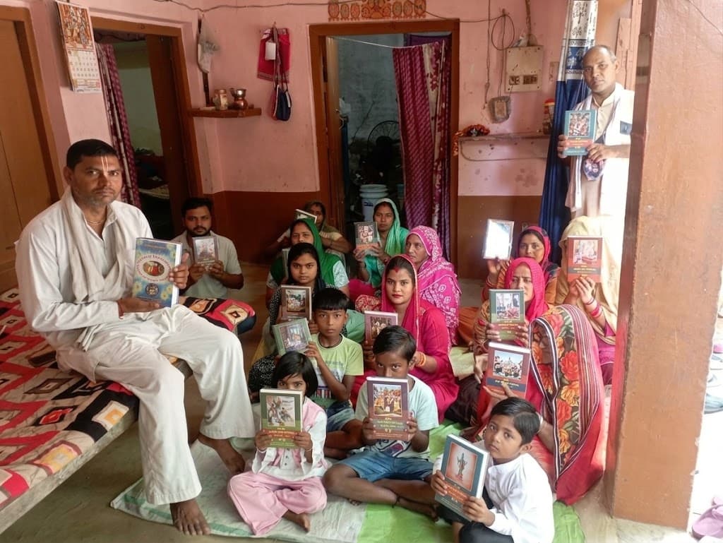 A village family and neighbors gathered in the doorway of their home, each holding a Śrīmad-Bhāgavatam volume