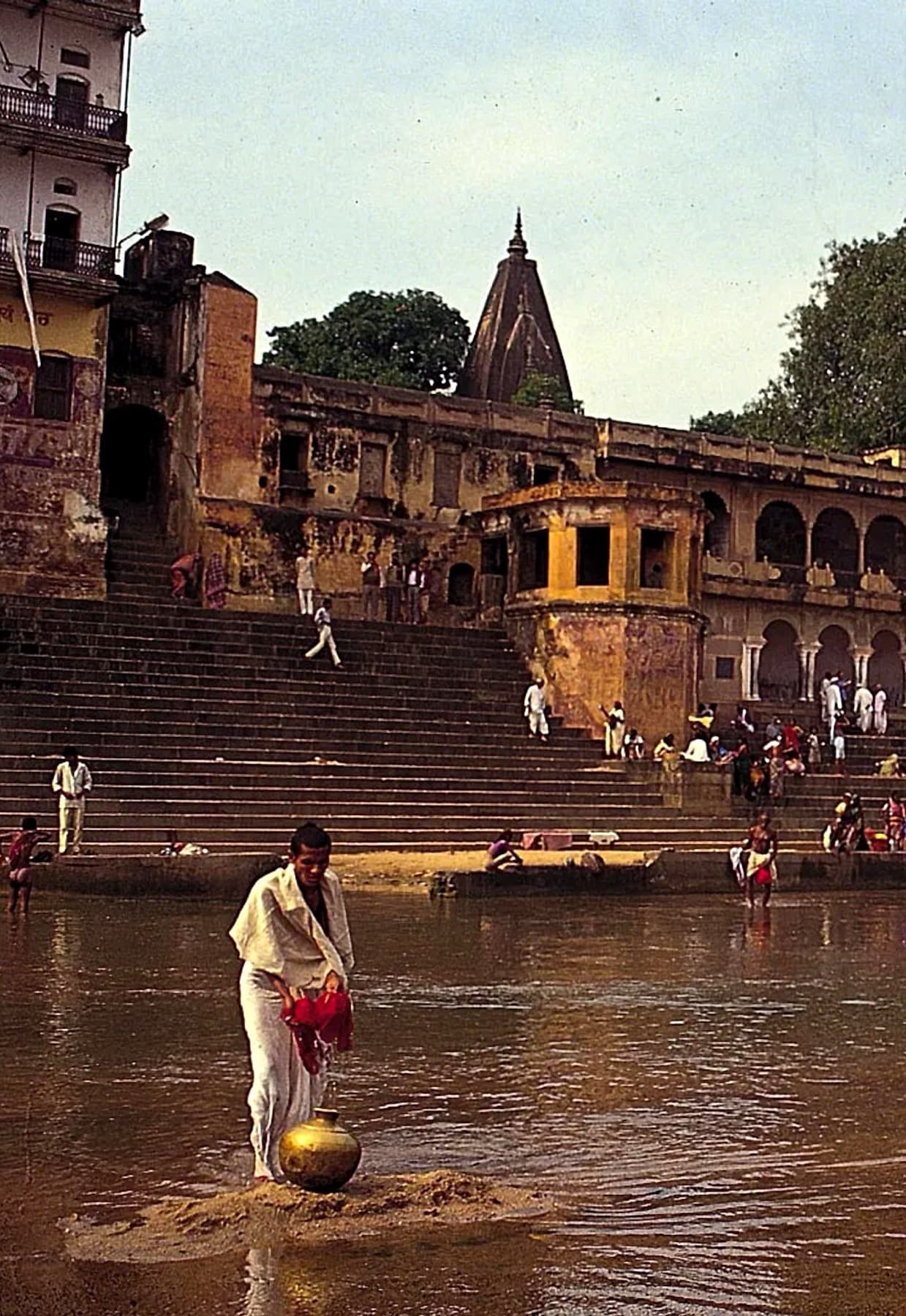 Pilgrims on the ghats of the Phalgu riverbank, a man in white carrying a brass vessel in the shallows